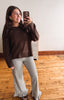 A woman takes a mirror selfie indoors wearing a dark brown knit sweater and black-and-white gingham flared pants with ruffled hems.