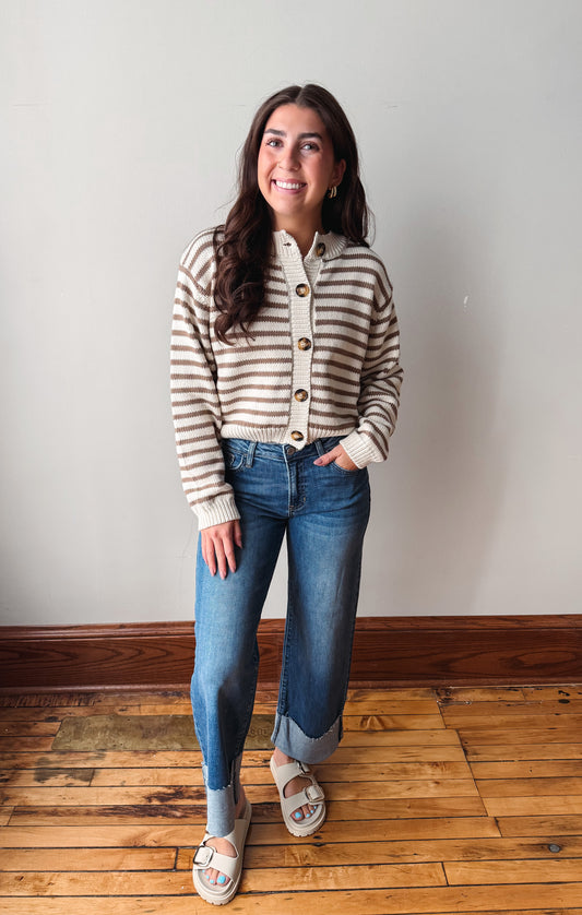 A woman stands in a room with a light gray wall and wooden floor, wearing a beige and brown striped button-up cardigan, blue jeans, and light-colored slide sandals.