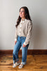 A woman modeling a cream and brown striped knit cardigan with wooden buttons, paired with blue jeans and white slide sandals, standing in a room with a light gray wall and wooden floor.