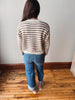A woman viewed from behind wearing a cream and brown horizontally striped knit sweater, blue jeans, and pink slip-on shoes, standing on a wooden floor against a plain light gray wall.