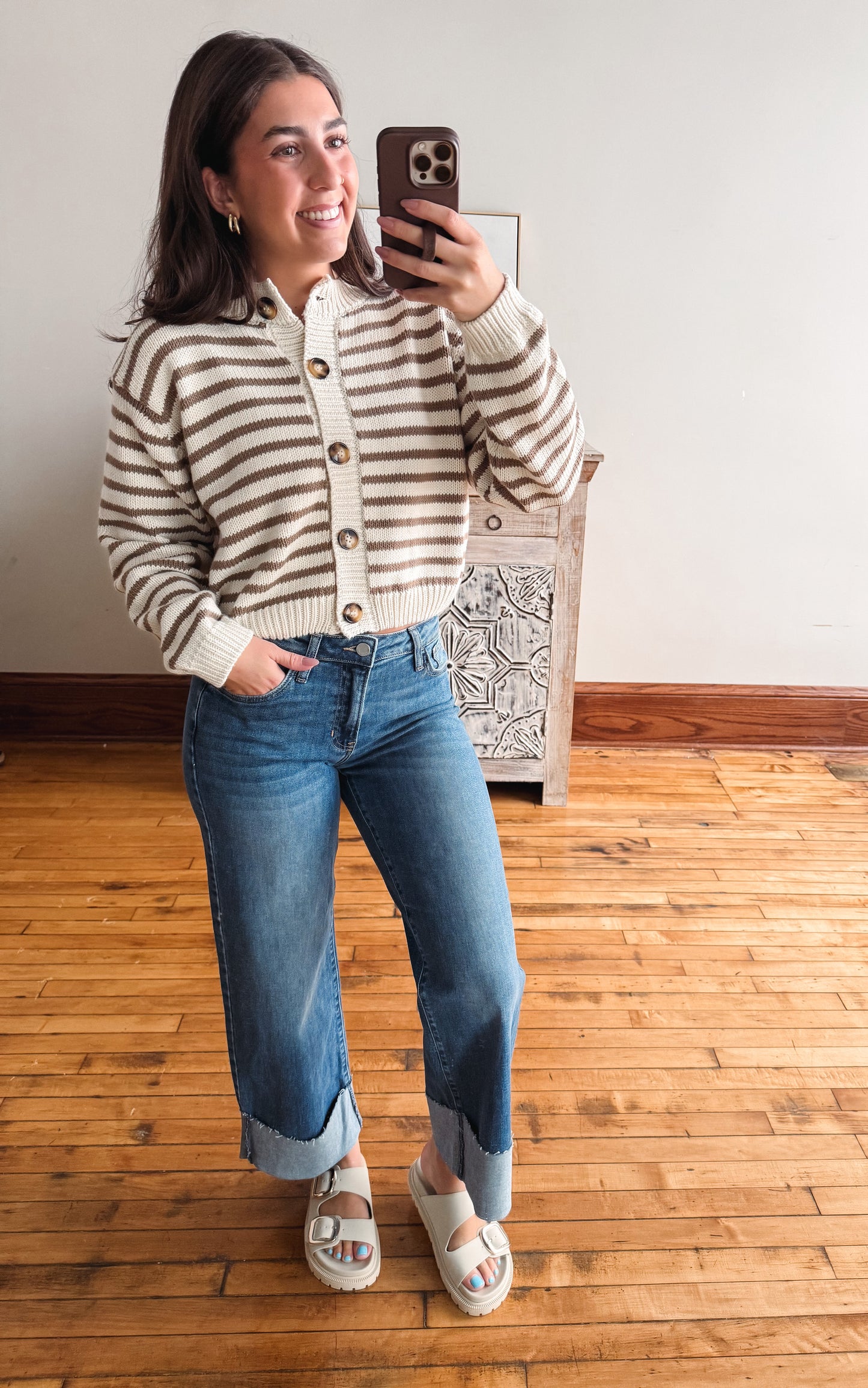 A woman takes a mirror selfie wearing a cream and brown horizontally striped knit cardigan, blue cuffed jeans, and white slide sandals in a room with a wooden floor and white wall.