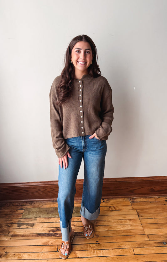 A woman stands in a room with a light gray wall and wooden floor, wearing a olive button-front knit cardigan, blue jeans, and brown sandals.