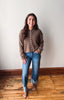 A woman stands in a room with a light gray wall and wooden floor, wearing a olive button-front knit cardigan, blue jeans, and brown sandals.