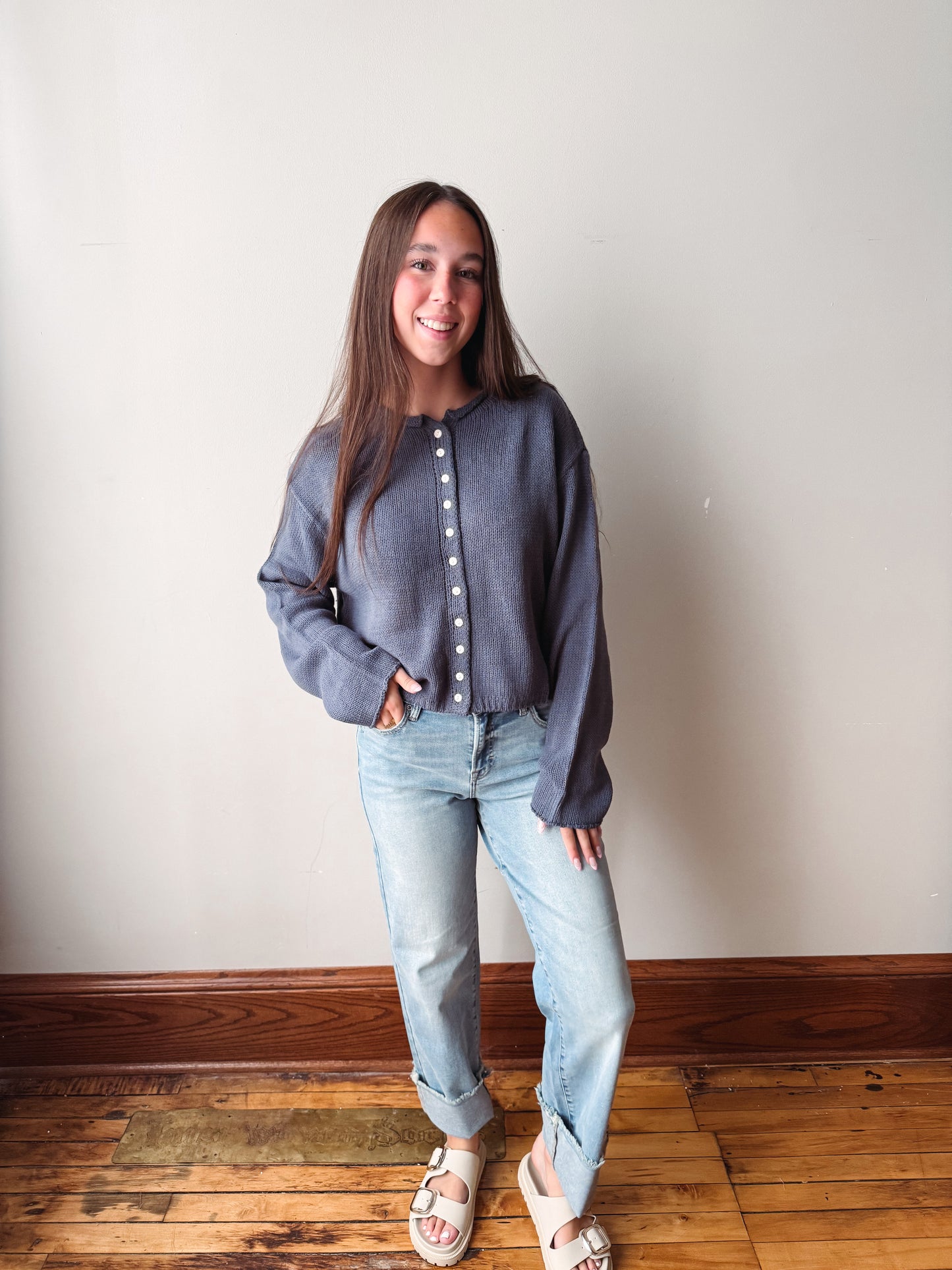 A young woman stands in a room with a light-colored wall and wooden floor, wearing a slate blue-gray long-sleeve button-up knit top, light-wash blue jeans, and white slide sandals.