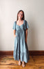 A woman modeling a dusty blue maxi dress with short flutter sleeves, a gathered sweetheart neckline, and side pockets, standing on a wooden floor against a plain beige wall.