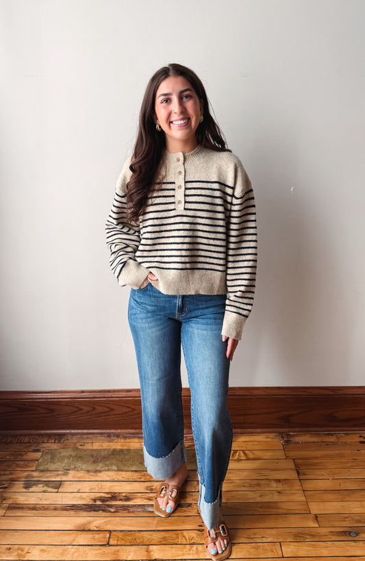 A woman stands in a room with a light gray wall and wooden floor, wearing a beige and black horizontally striped knit sweater, blue jeans, and brown sandals.