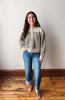 A woman stands in a room with a light gray wall and wooden floor, wearing a beige and black horizontally striped knit sweater, blue jeans, and brown sandals.