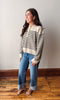 A woman wearing a cream and black striped knit sweater, blue jeans, and brown sandals, standing on a wooden floor against a plain light gray wall.