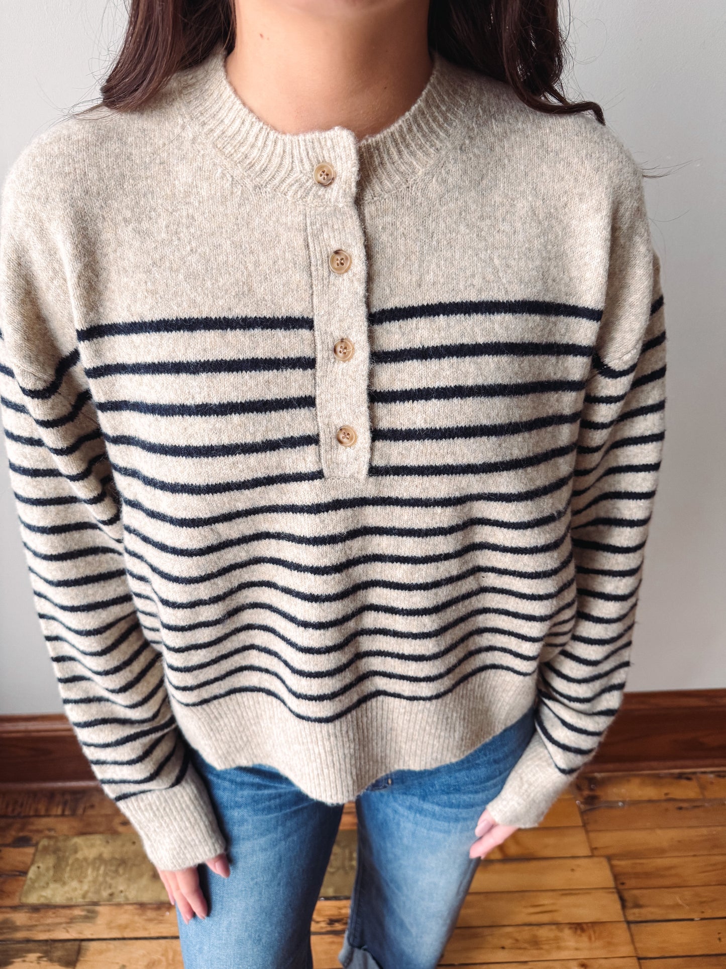 A person wearing a cream knit henley sweater with navy horizontal stripes and four wooden buttons, paired with blue jeans, standing on a wooden floor against a plain wall.