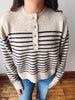 A person wearing a cream knit henley sweater with navy horizontal stripes and four wooden buttons, paired with blue jeans, standing on a wooden floor against a plain wall.