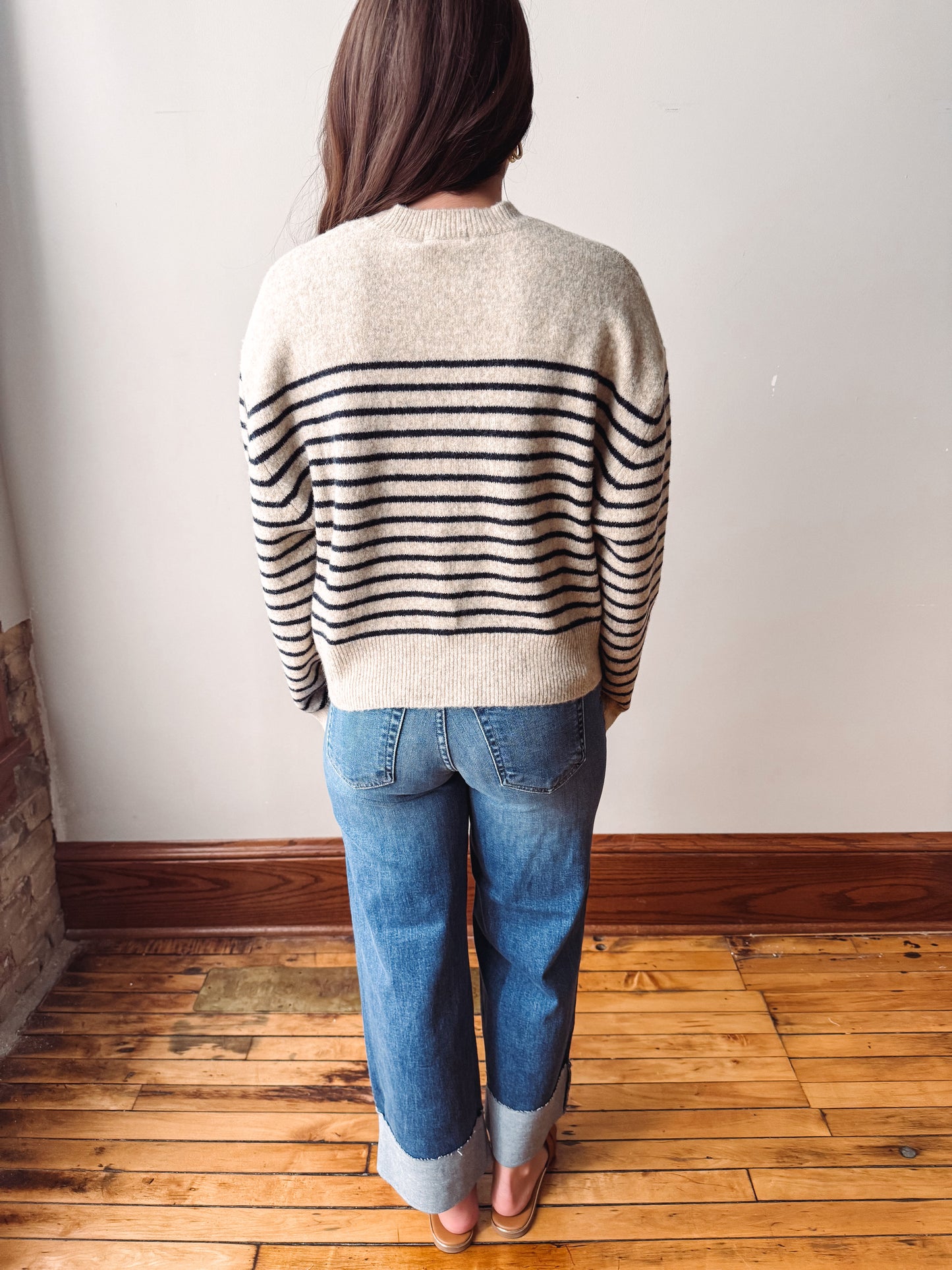A woman viewed from behind wearing a cream knit sweater with black horizontal stripes on the sleeves and back, paired with blue jeans and tan flats, standing on a wooden floor against a white wall.