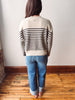 A woman viewed from behind wearing a cream knit sweater with black horizontal stripes on the sleeves and back, paired with blue jeans and tan flats, standing on a wooden floor against a white wall.