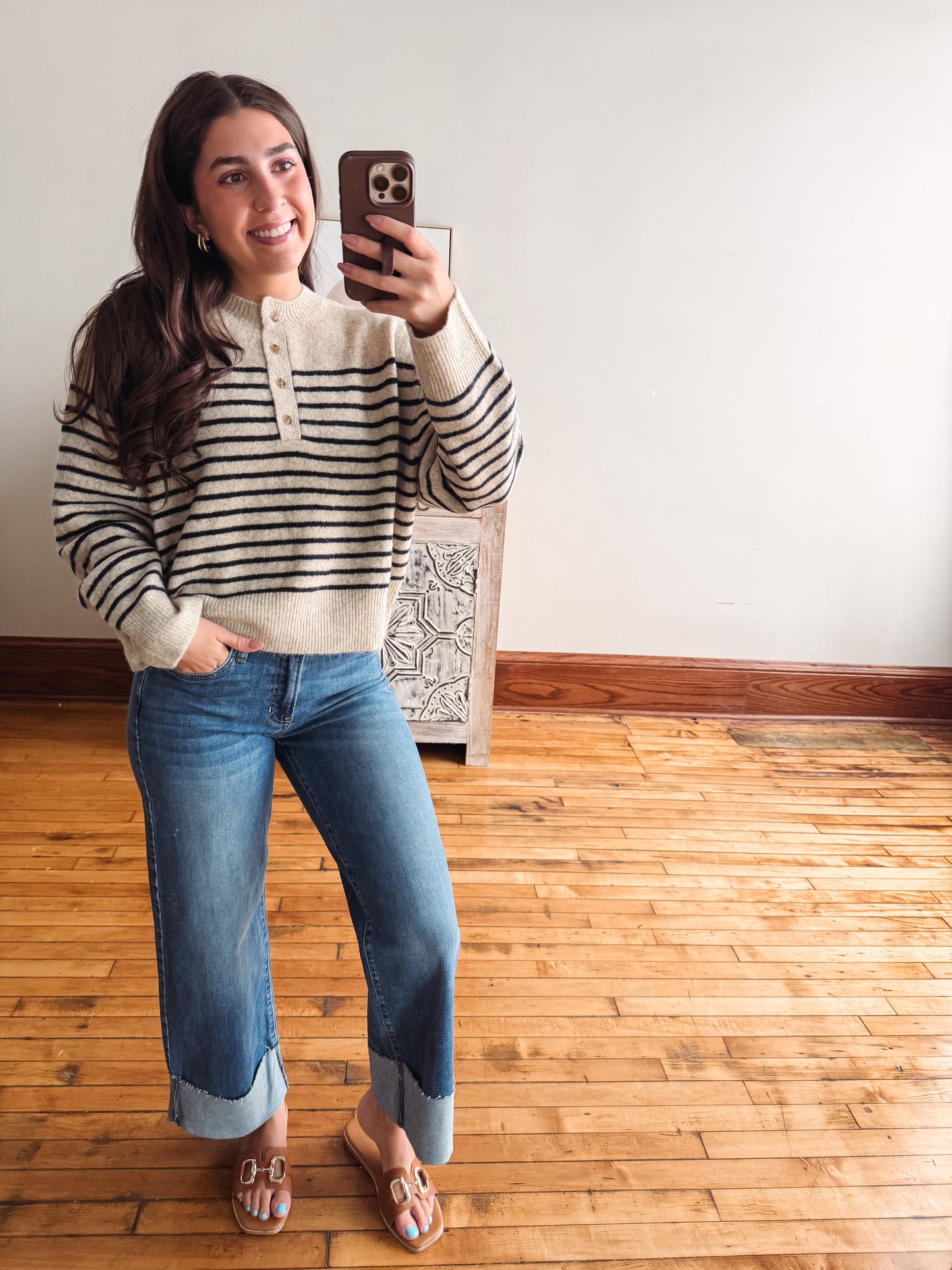 A woman takes a mirror selfie wearing a cream and navy striped long-sleeve henley sweater, blue cuffed jeans, and tan slide sandals in a room with light wood floors and a white wall.