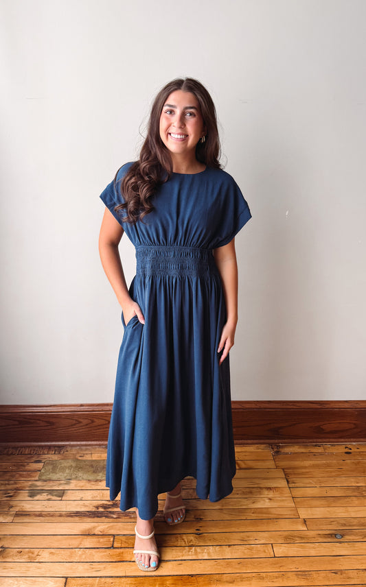 A woman modeling a navy blue maxi dress with short flutter sleeves, a smocked waist, and side pockets, standing on a wooden floor against a plain off-white wall.