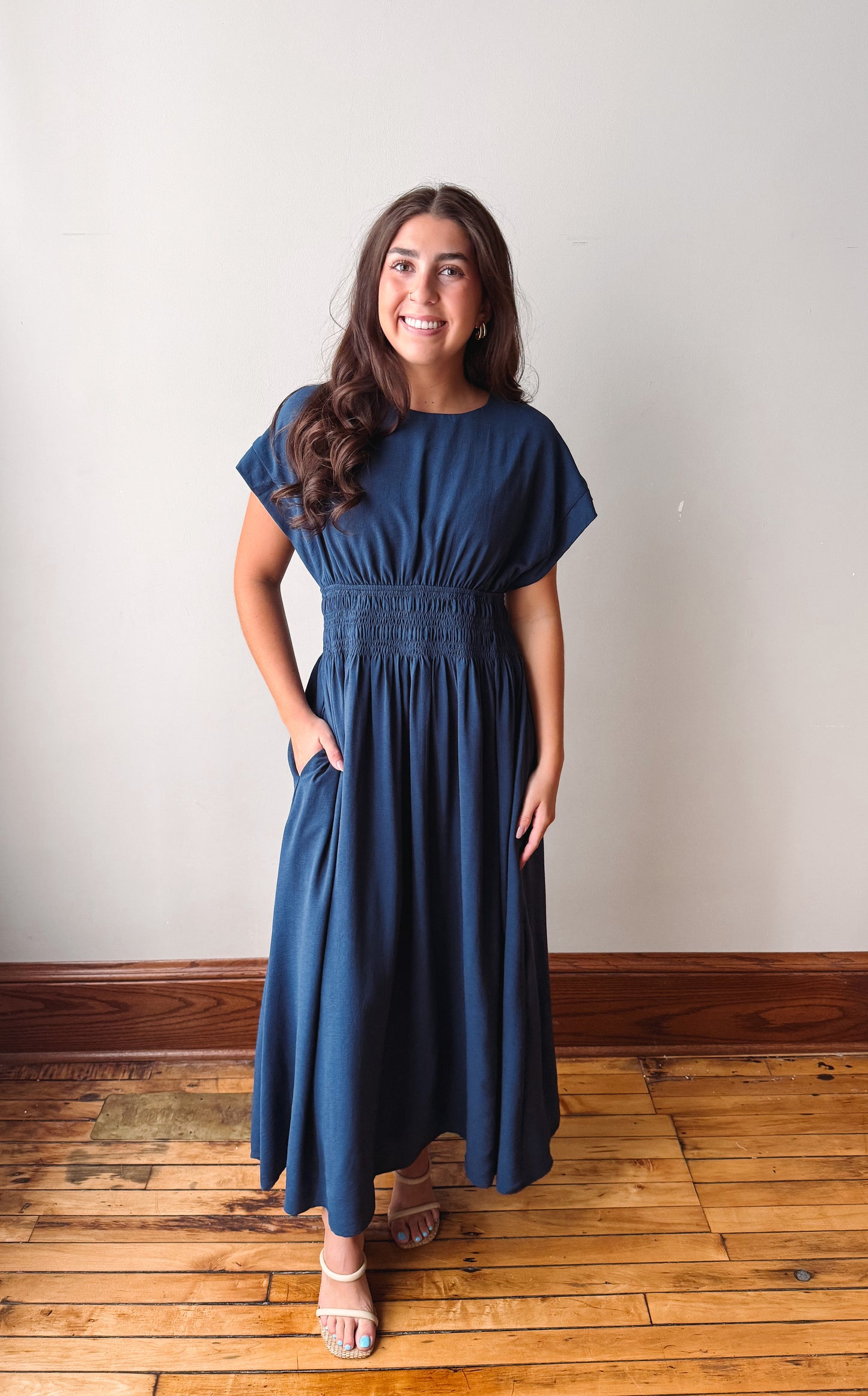 A woman modeling a navy blue maxi dress with short flutter sleeves, a smocked waist, and side pockets, standing on a wooden floor against a plain off-white wall.