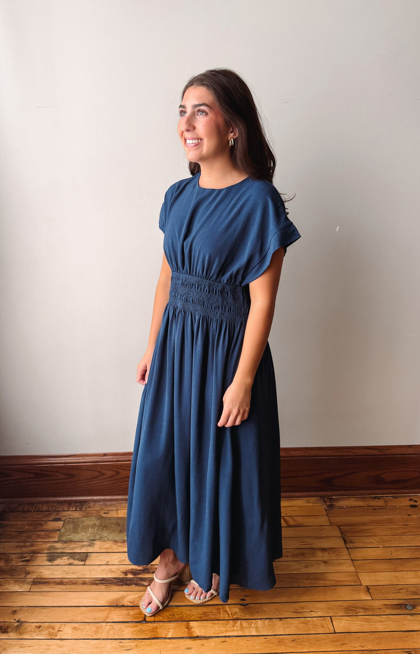 A woman modeling a navy blue maxi dress with flutter sleeves and a smocked waist, standing on a wooden floor against a plain off-white wall.