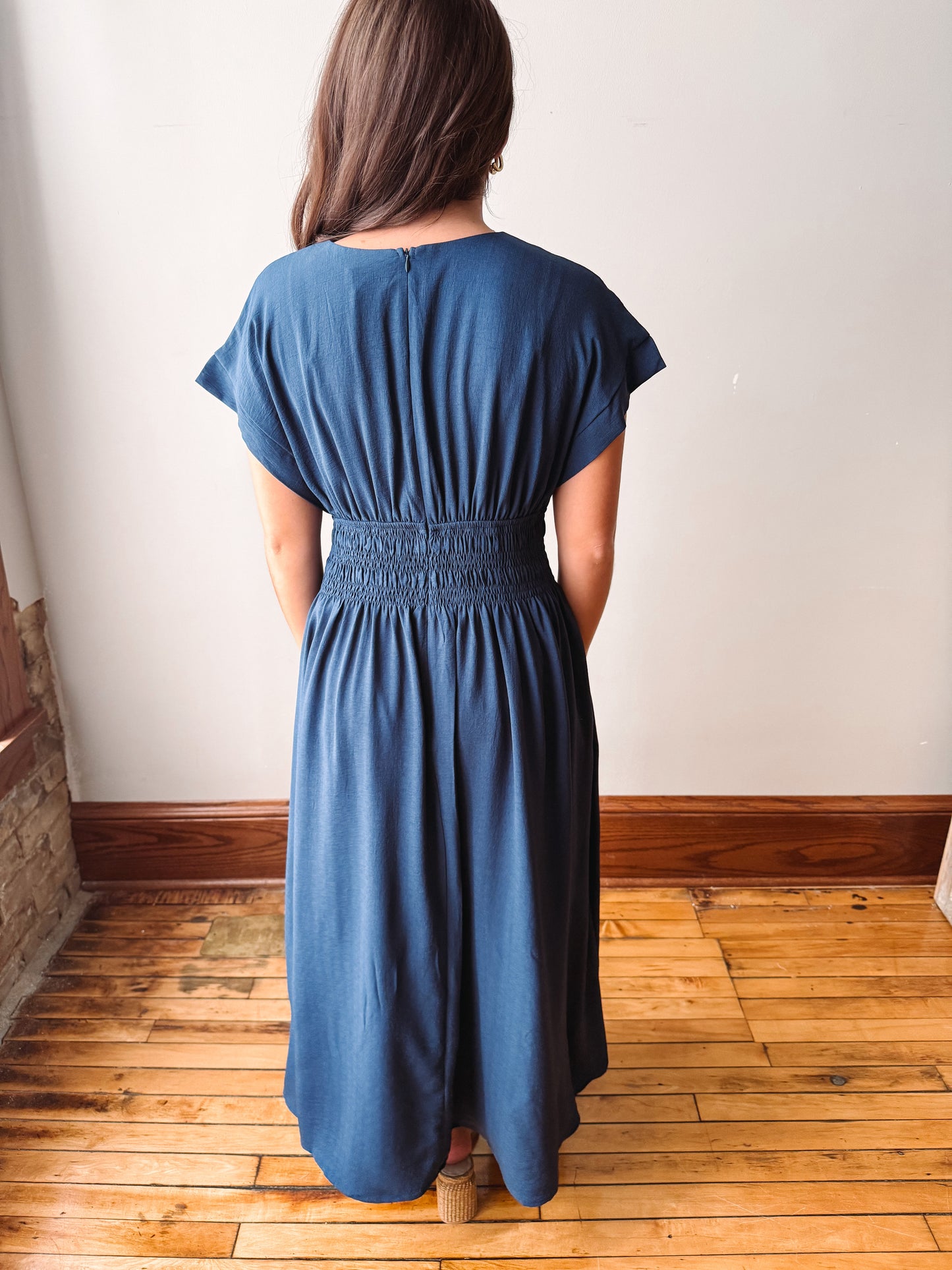 A woman viewed from behind wearing a short-sleeve blue midi dress with a smocked waist, standing on a wooden floor against a plain wall.