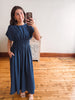 A woman takes a mirror selfie wearing a navy blue maxi dress with short flutter sleeves, a smocked waist, and a gathered skirt, standing in a room with wooden floors and a white wall.