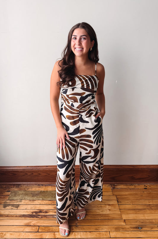 A woman modeling a sleeveless wide-leg jumpsuit with a brown, black, and beige palm leaf pattern, standing on a wooden floor against a plain light gray wall.