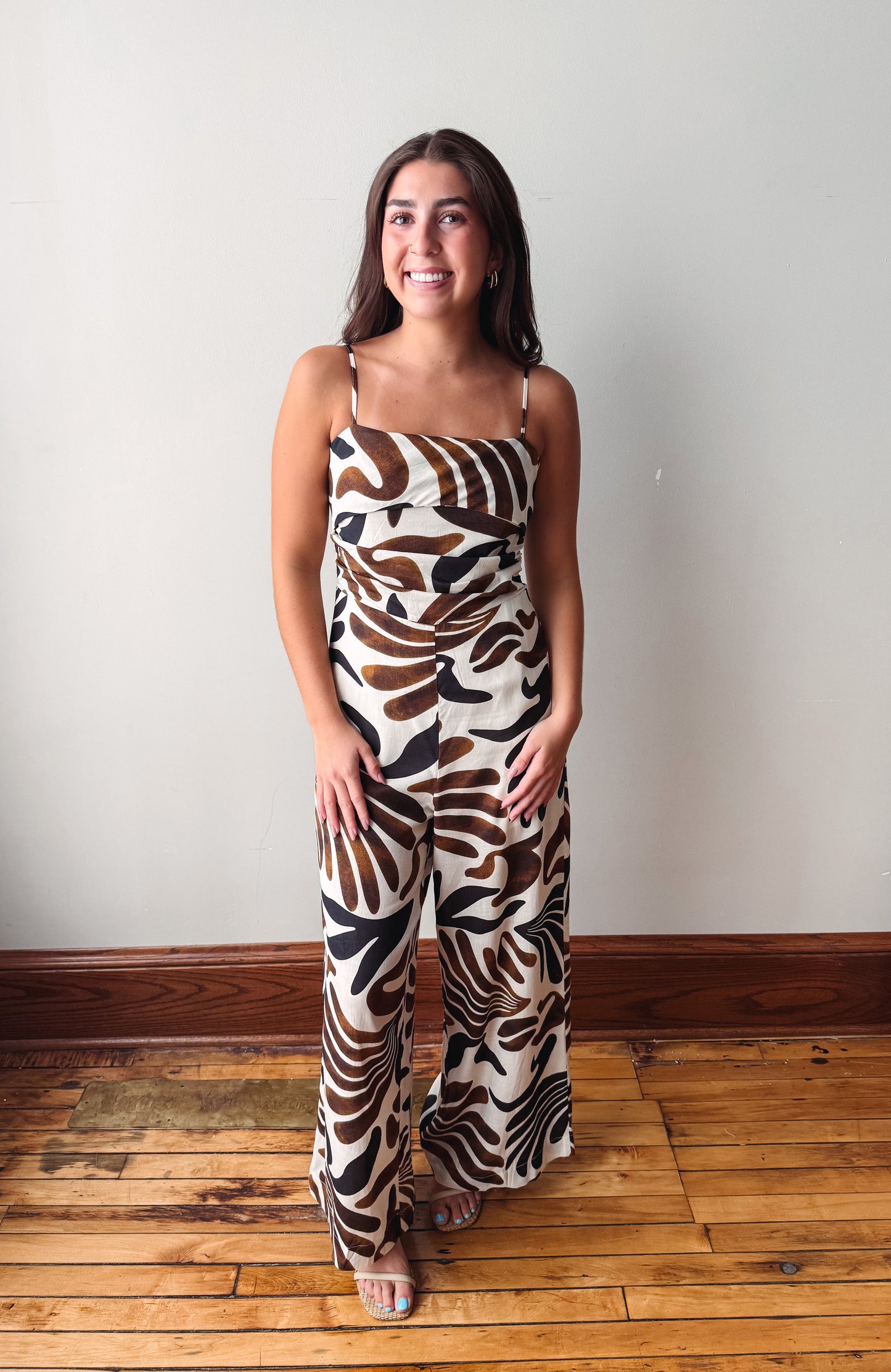 A woman modeling a sleeveless wide-leg jumpsuit with a brown, black, and cream abstract leaf pattern, standing on a wooden floor against a plain light gray wall.