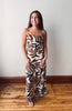 A woman modeling a sleeveless wide-leg jumpsuit with a brown, black, and cream abstract leaf pattern, standing on a wooden floor against a plain light gray wall.