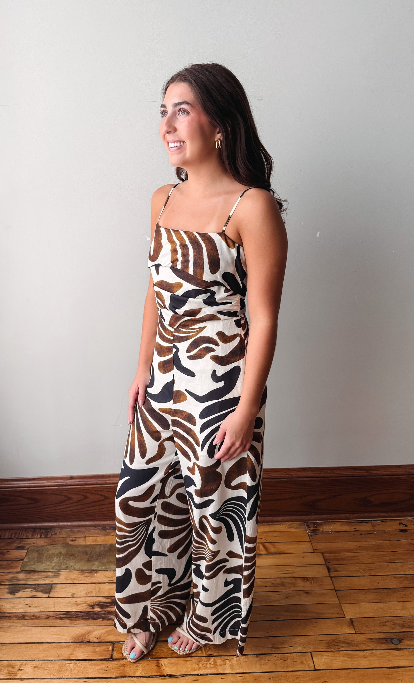 A woman modeling a sleeveless wide-leg jumpsuit with a brown, black, and cream abstract animal-stripe pattern, standing on a wooden floor against a plain light gray wall.