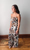 A woman modeling a sleeveless wide-leg jumpsuit with a brown, black, and cream abstract animal-stripe pattern, standing on a wooden floor against a plain light gray wall.