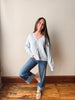 A woman posing in a light blue button-up cardigan, blue jeans, and beige sandals against a beige wall and wooden floor.