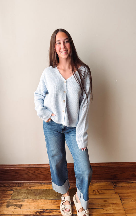 A woman stands indoors wearing a light blue fuzzy button-front cardigan, blue jeans, and white slide sandals against a plain beige wall and wood floor.