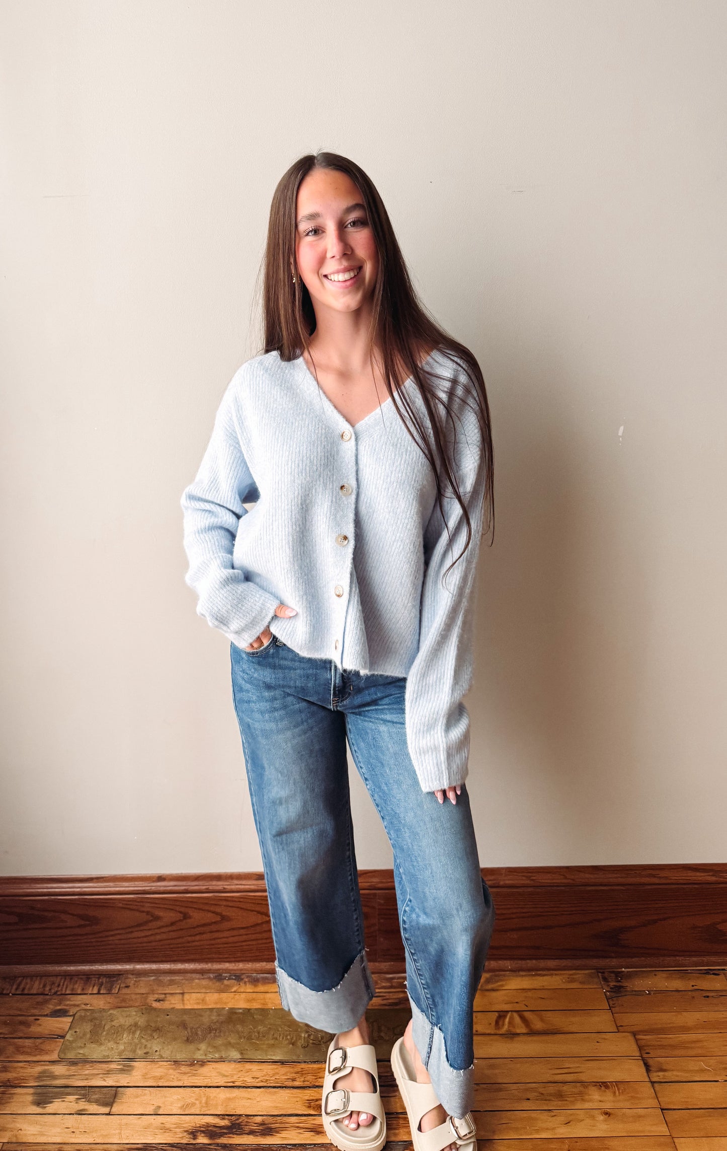 A woman stands indoors wearing a light blue fuzzy button-front cardigan, blue jeans, and white slide sandals against a plain beige wall and wood floor.