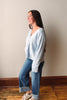 A woman wearing a light blue knit cardigan, blue jeans, and beige sandals, standing in profile against a beige wall with a wooden baseboard and wood floor.