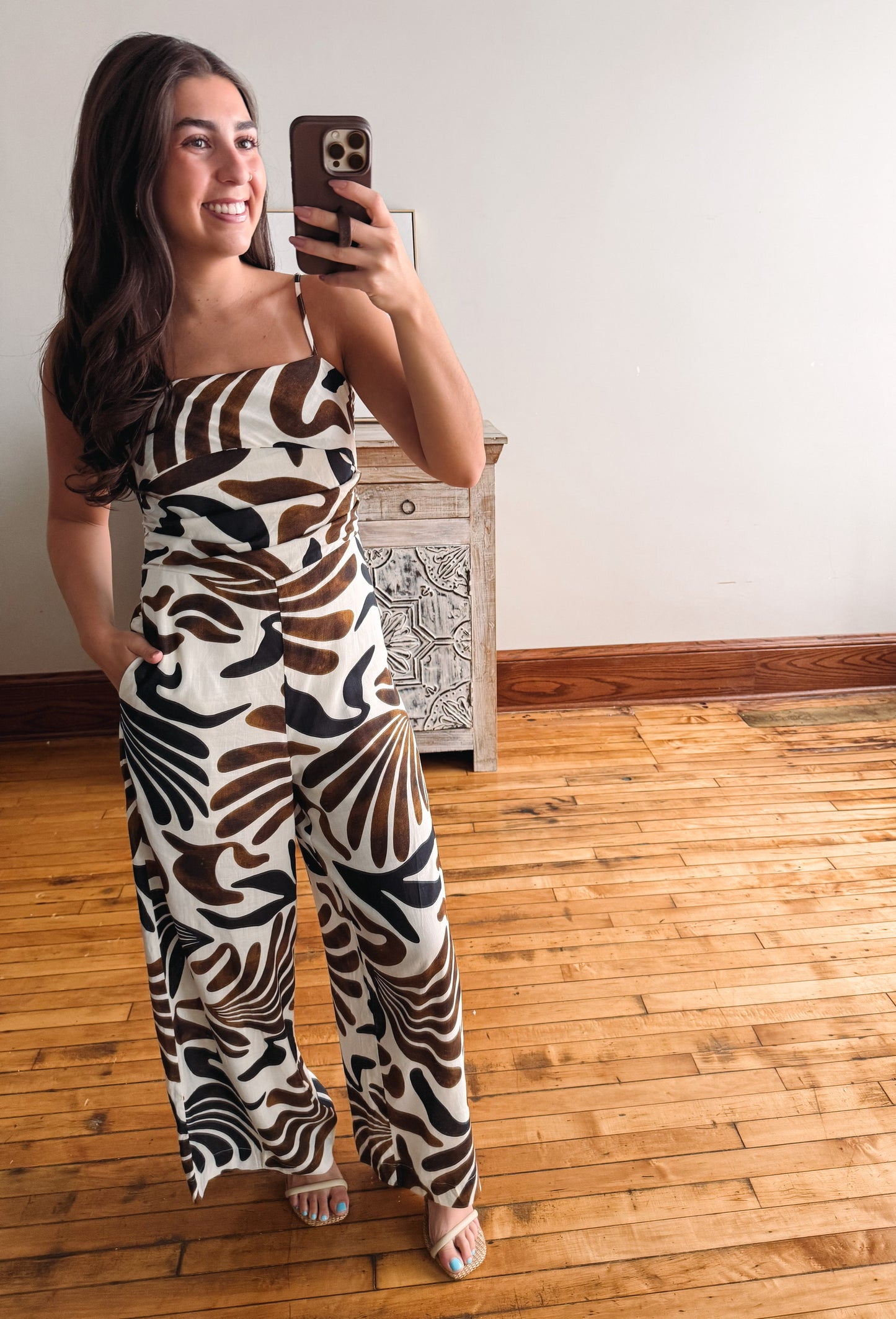 A woman takes a mirror selfie wearing a strapless jumpsuit with a bold brown, black, and white abstract pattern. She is standing on a light wood floor in front of a white wall and a small wooden cabinet.
