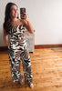 A woman takes a mirror selfie wearing a strapless jumpsuit with a bold brown, black, and white abstract pattern. She is standing on a light wood floor in front of a white wall and a small wooden cabinet.