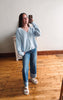 A woman takes a mirror selfie wearing a light blue knit cardigan, blue jeans, and white slide sandals in a room with a wooden floor and off-white wall.