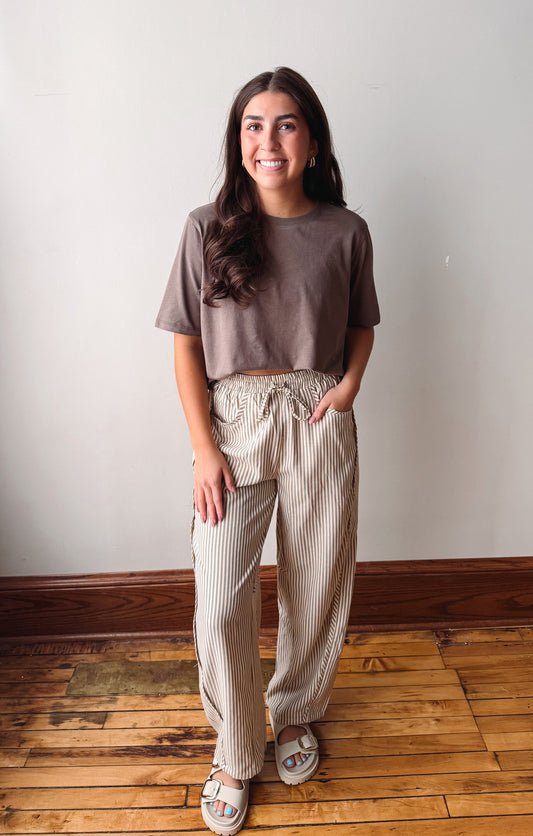 A woman stands indoors wearing a taupe short-sleeve crop top and beige-and-white vertically striped wide-leg pants, paired with white slide sandals, against a plain off-white wall and wooden floor.
