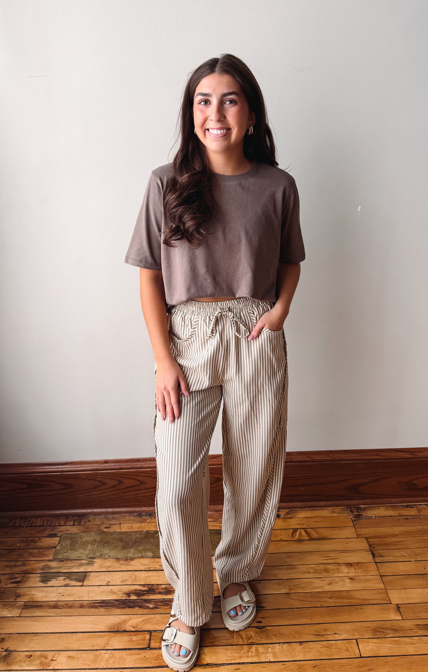 A woman stands indoors wearing a taupe short-sleeve crop top and beige-and-white vertically striped wide-leg pants, paired with white slide sandals, against a plain off-white wall and wooden floor.