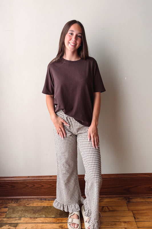 A woman stands indoors against a plain off-white wall, wearing a dark brown short-sleeve t-shirt and beige-and-black gingham wide-leg ruffled pants, paired with white sandals.