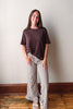 A woman stands indoors against a plain off-white wall, wearing a dark brown short-sleeve t-shirt and beige-and-black gingham wide-leg ruffled pants, paired with white sandals.