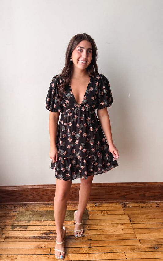 A woman modeling a short black dress with a pink and green floral pattern, featuring puff sleeves and a ruffled hem, standing on a wooden floor against a plain wall.