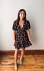 A woman modeling a short black dress with a pink and green floral pattern, featuring puff sleeves and a ruffled hem, standing on a wooden floor against a plain wall.
