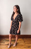A woman modeling a short black dress with a pink and green tropical floral print, featuring puff sleeves and a deep V-neckline, standing on a wooden floor against a plain wall.