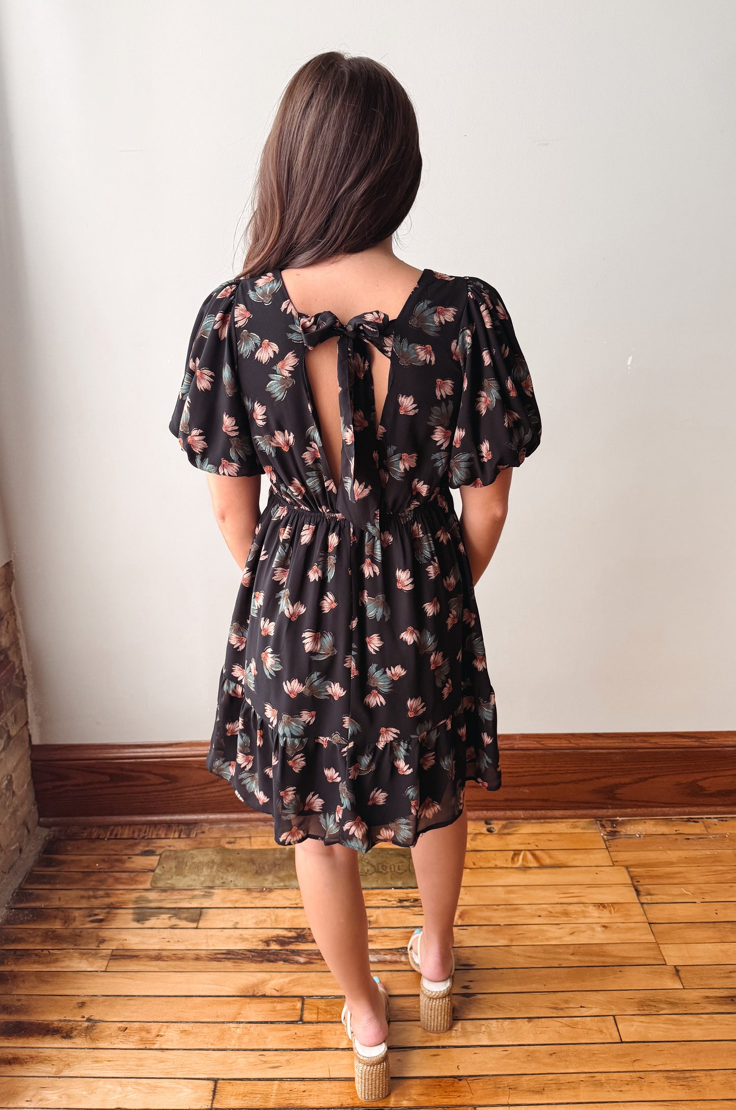 A woman viewed from behind wearing a black short-sleeve dress with a pink and green floral pattern, puff sleeves, and a tie detail at the back neckline. She is standing on a wooden floor against a plain off-white wall.