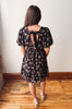 A woman viewed from behind wearing a black short-sleeve dress with a pink and green floral pattern, puff sleeves, and a tie detail at the back neckline. She is standing on a wooden floor against a plain off-white wall.