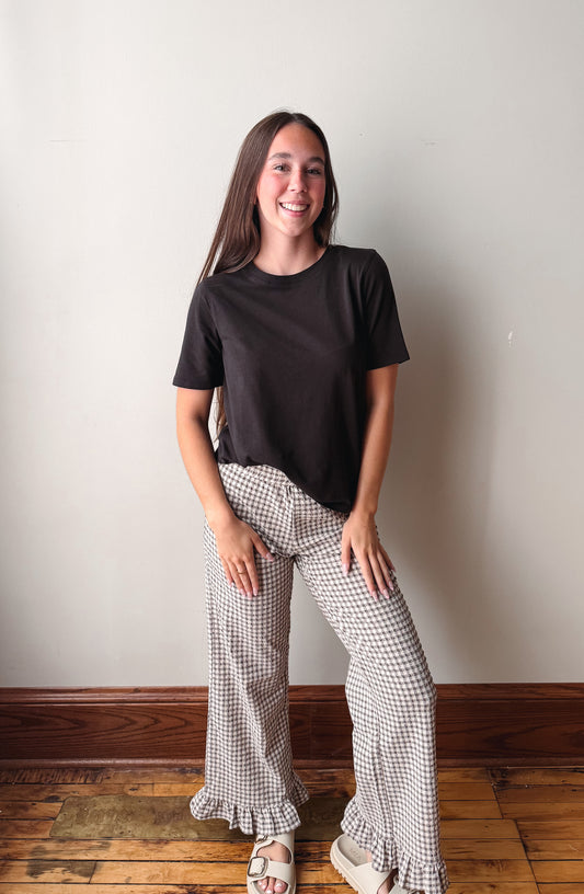A young woman stands in a room with a light gray wall and wooden floor, wearing a black short-sleeve t-shirt and beige-and-black checkered wide-leg ruffled pants.