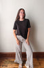 A young woman stands in a room with a light gray wall and wooden floor, wearing a black short-sleeve t-shirt and beige-and-black checkered wide-leg ruffled pants.