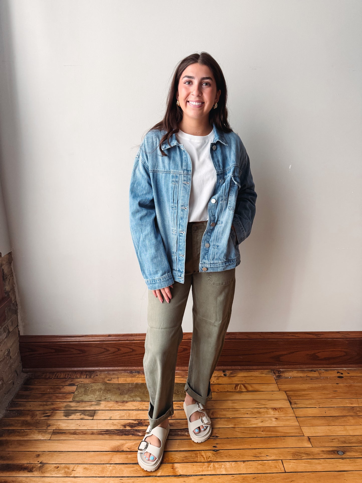 A woman stands indoors wearing a light blue denim jacket over a white top, olive green pants, and white slide sandals, photographed against a plain off-white wall with a wood floor.