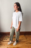 A woman modeling a white short-sleeve crewneck t-shirt, paired with olive green pants and white slide sandals, standing on a wooden floor against a plain wall.