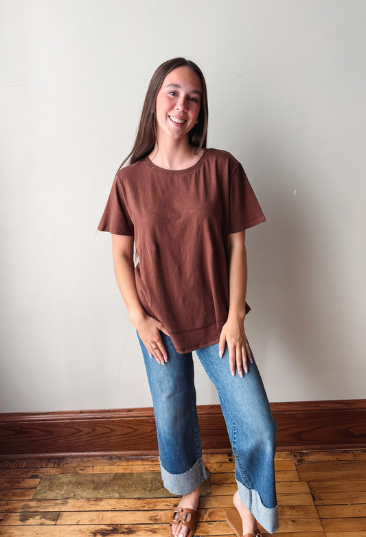 A young woman smiling while wearing a brown short-sleeve t-shirt and blue jeans with cuffed hems, standing on a wooden floor against a plain off-white wall.