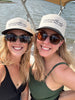 Two women smiling and taking a selfie on a boat, wearing matching off-white trucker hats with black brims and the text 'URBANthreads CLOTHING BOUTIQUE' printed on the front.