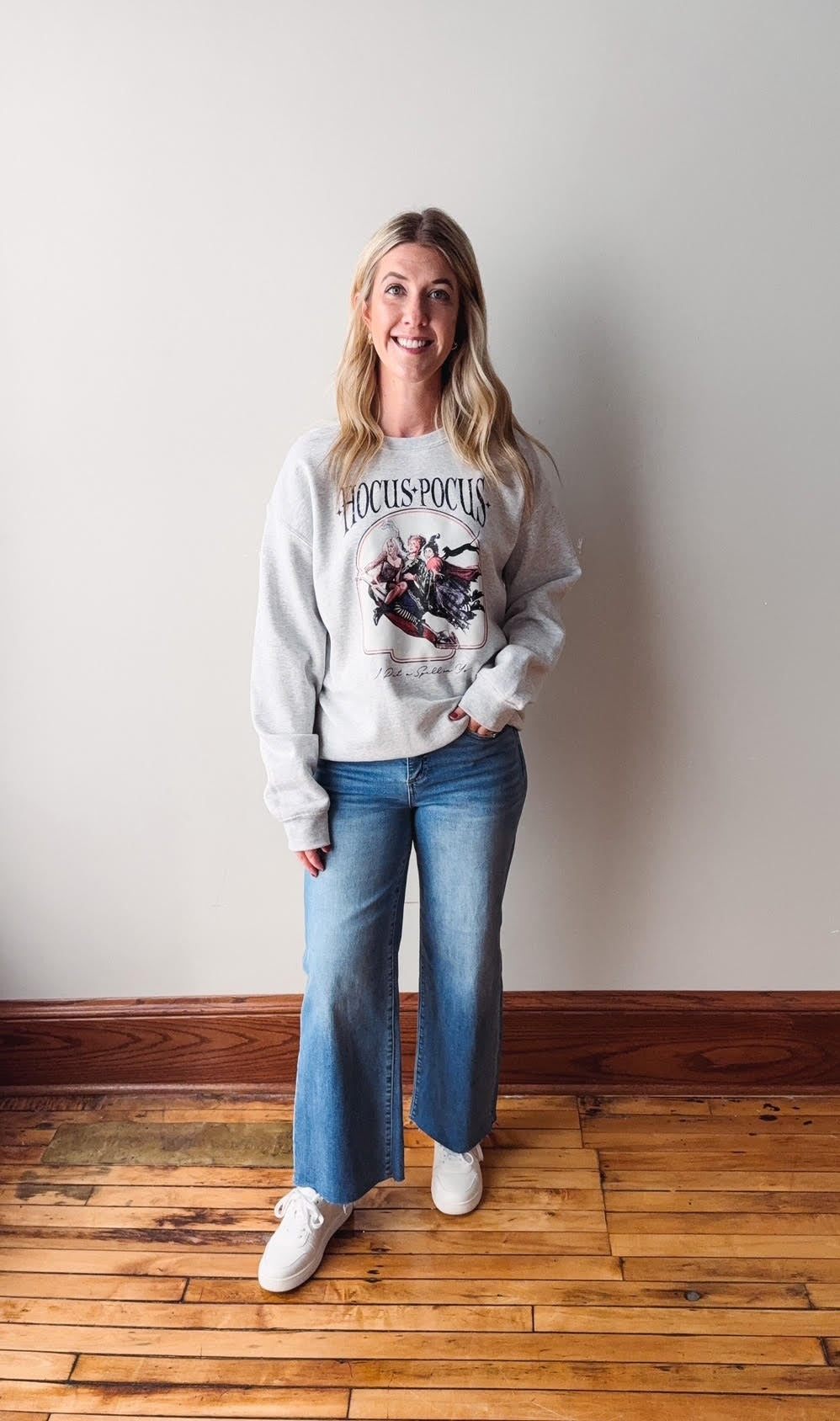 Woman wearing a white sweatshirt with graphic design and blue jeans standing against a plain wall.